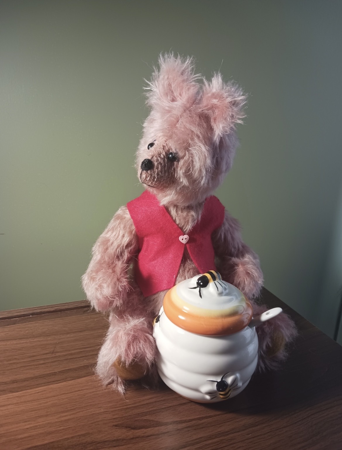 Handmade pink bear wearing a red vest beside a honey pot